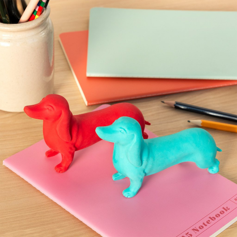 Zwei Dackel-Radiergummis in den Farben Rot und Türkis stehen auf einem pinken Notizbuch. Im Hintergrund liegen Stifte und Hefte auf einem Holzschreibtisch.