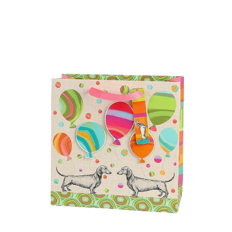 Bunte Geschenktasche mit Dackel-Illustrationen, farbenfrohen Ballons und rosa Trageschlaufe, verziert mit verspieltem Punkt- und Kreismuster.