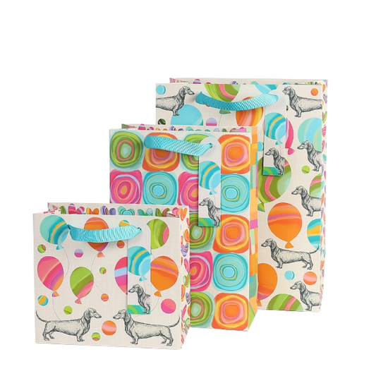 Drei bunte Geschenktaschen mit Dackel-Illustrationen und Ballon-Motiven in verschiedenen Größen, jede mit blauer Trageschlaufe.