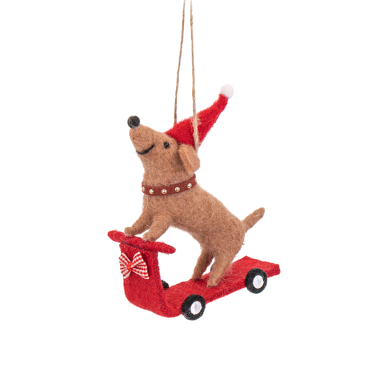 Weihnachtlicher Filzanhänger eines Dackels mit roter Mütze auf einem roten Roller mit Schleife.