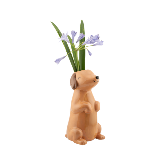 vase in Form eines sitzenden, fröhlich dreinblickenden Dackels. Aus dem Kopf der Figur ragen lila Blüten und grüne Blumenstängel wie aus einer klassischen Vase.