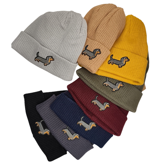 Mehrere bunte Strickmützen in verschiedenen Farben wie Grau, Senfgelb, Schwarz, Bordeaux und Olivgrün, jede mit einem aufgestickten kleinen Dackel auf dem umgeschlagenen Rand.