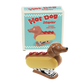 Witziger Klammerhefter im Stil eines Hot Dogs mit Dackelkopf. Der Hefter hat einen gelben „Brötchen“-Körper, eine rote Ketchup-Linie oben und ein süßes Dackelgesicht. Büroklammern liegen daneben.
