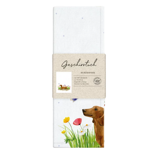 Weißes Geschirrtuch mit Aquarell-Illustration eines braunen Dackels, der durch eine bunte Blumenwiese läuft; im Vordergrund gelbe und rote Blüten sowie ein flatternder Schmetterling. Verpackt mit Papierbanderole.