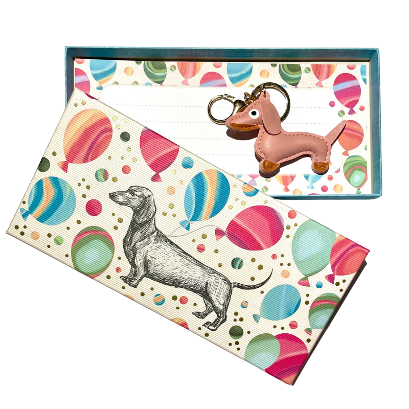 Geschenkbox mit buntem Ballonmuster und gezeichneter Dackel-Illustration, darin ein rosa Dackel-Schlüsselanhänger mit goldener Schlaufe auf gemustertem Hintergrund.