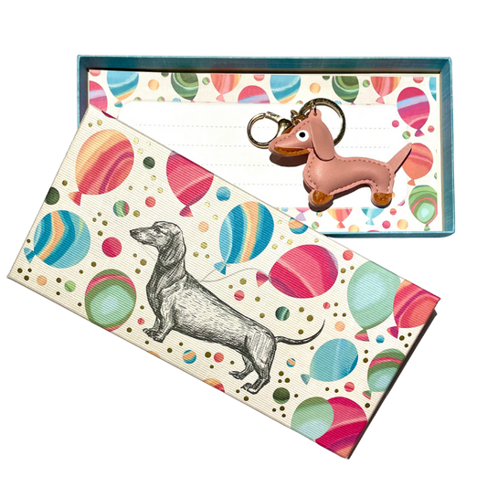 Geschenkbox mit buntem Ballonmuster und gezeichneter Dackel-Illustration, darin ein rosa Dackel-Schlüsselanhänger mit goldener Schlaufe auf gemustertem Hintergrund.