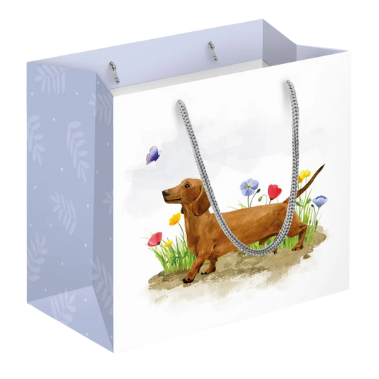 Geschenktasche mit frühlingshaftem Dackel-Motiv: brauner Dackel läuft durch ein buntes Blumenfeld, ein Schmetterling fliegt darüber, Seitenflächen in hellem Blau mit Blattmuster.
