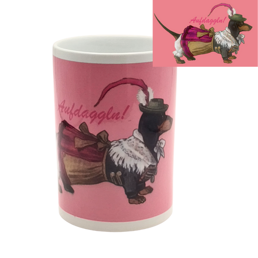 Weiße Tasse mit pinkem Hintergrund und Aufdruck eines Dackels in bayerischer Tracht, der ein Dirndl trägt und mit dem Spruch „Aufdaggl’n!“ versehen ist.